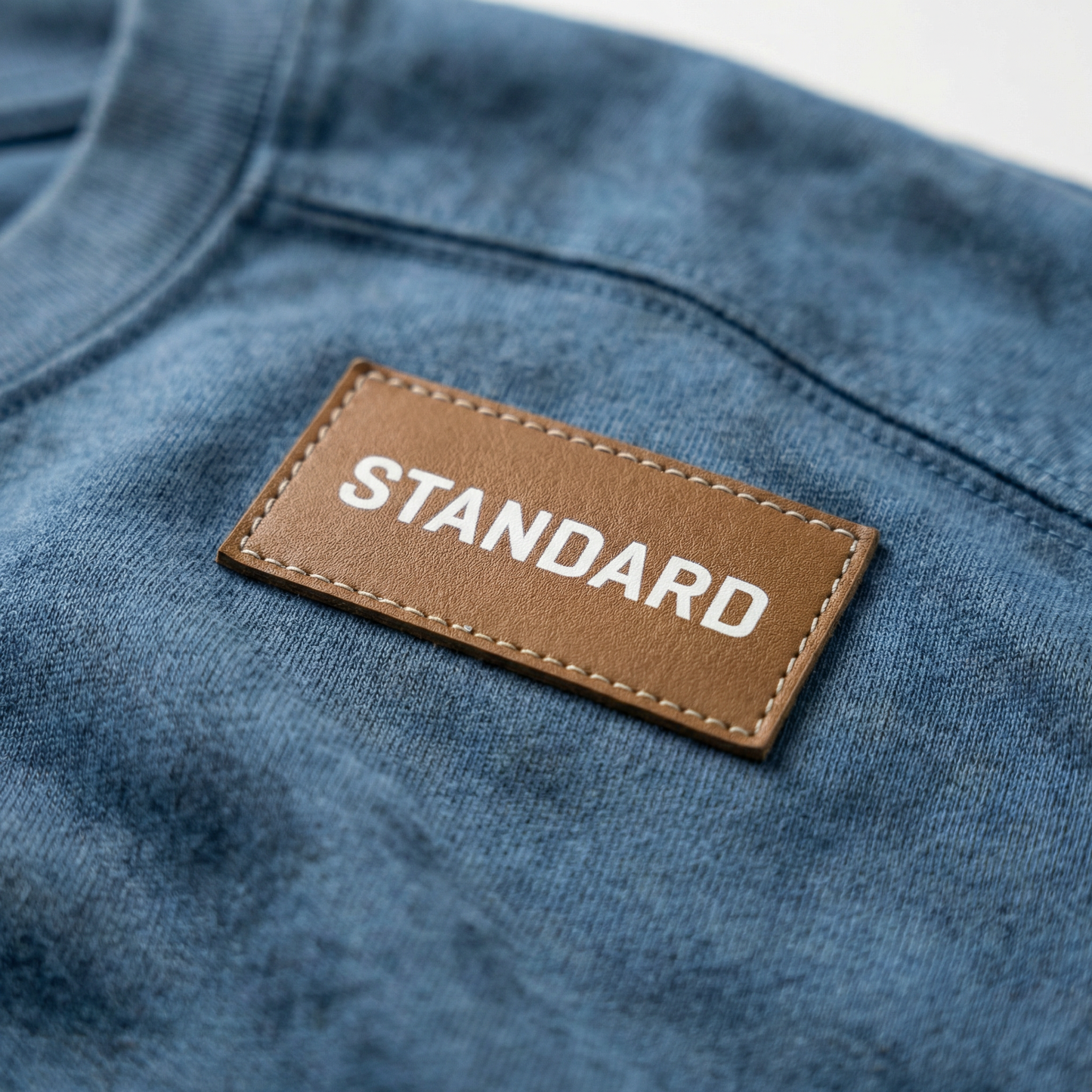 Macro detail of stitched leather shoulder patch and acid-wash effect on Blue cotton tee.
