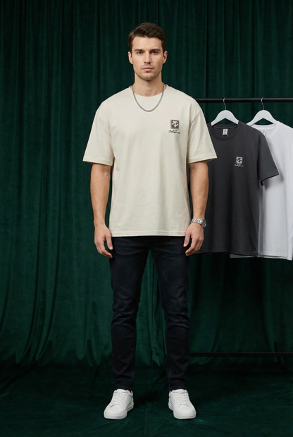 Man wearing WEARON Heritage Crest Tee in Ivory with hanging colourways behind, deep green velvet backdrop, full body.