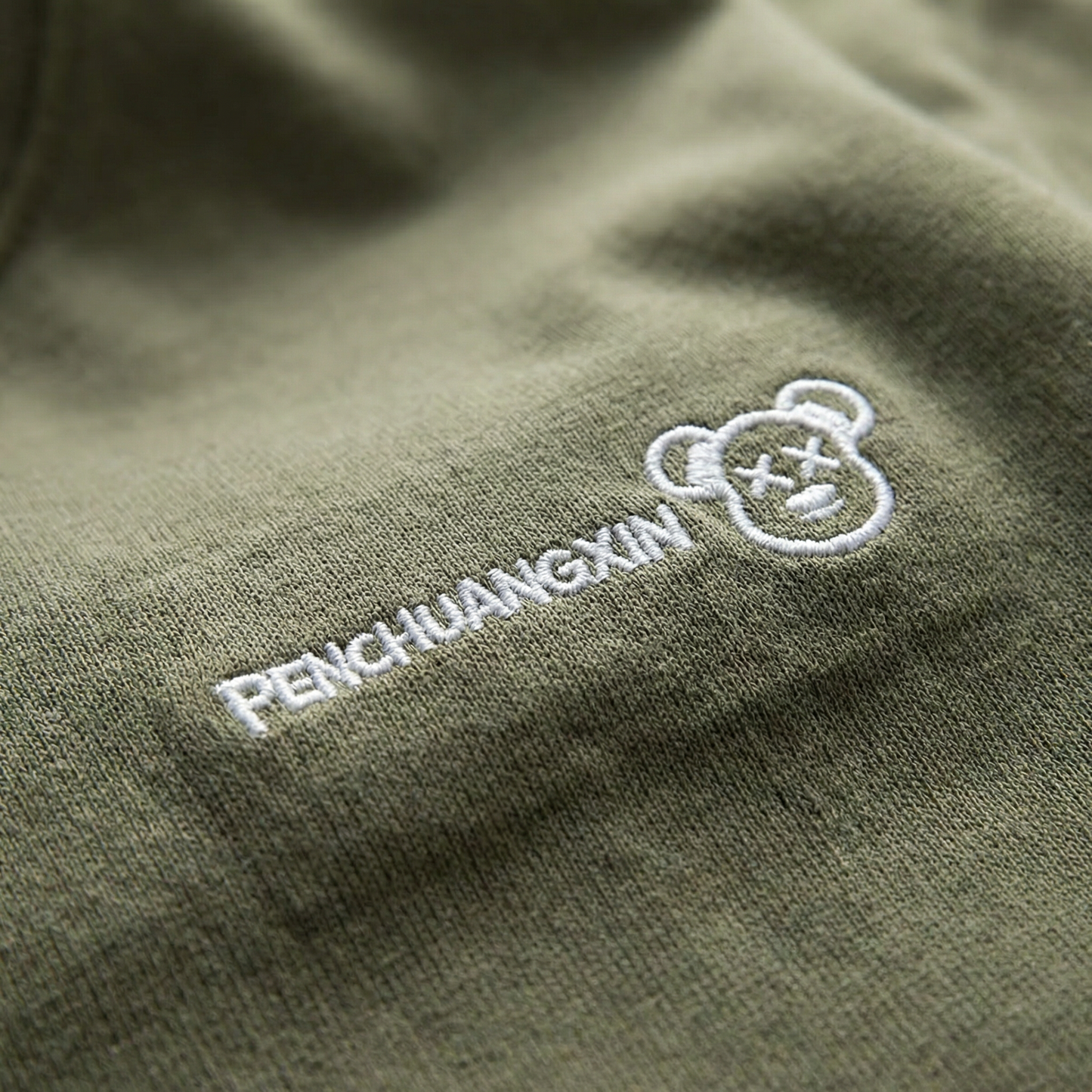 Macro detail of embroidered bear-face motif on Olive cotton tee.