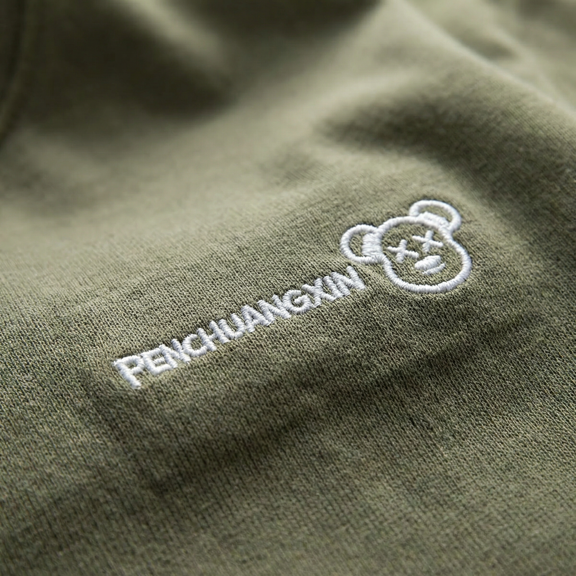 Macro detail of embroidered bear-face motif on Olive cotton tee.
