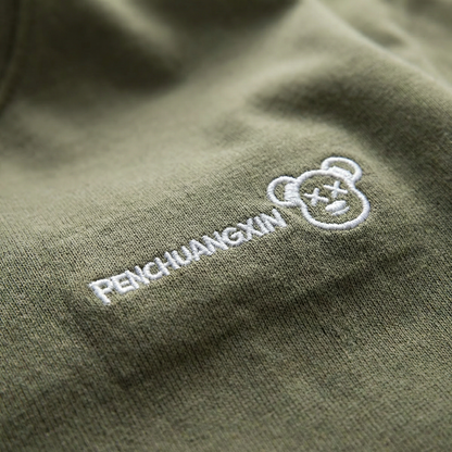 Macro detail of embroidered bear-face motif on Olive cotton tee.
