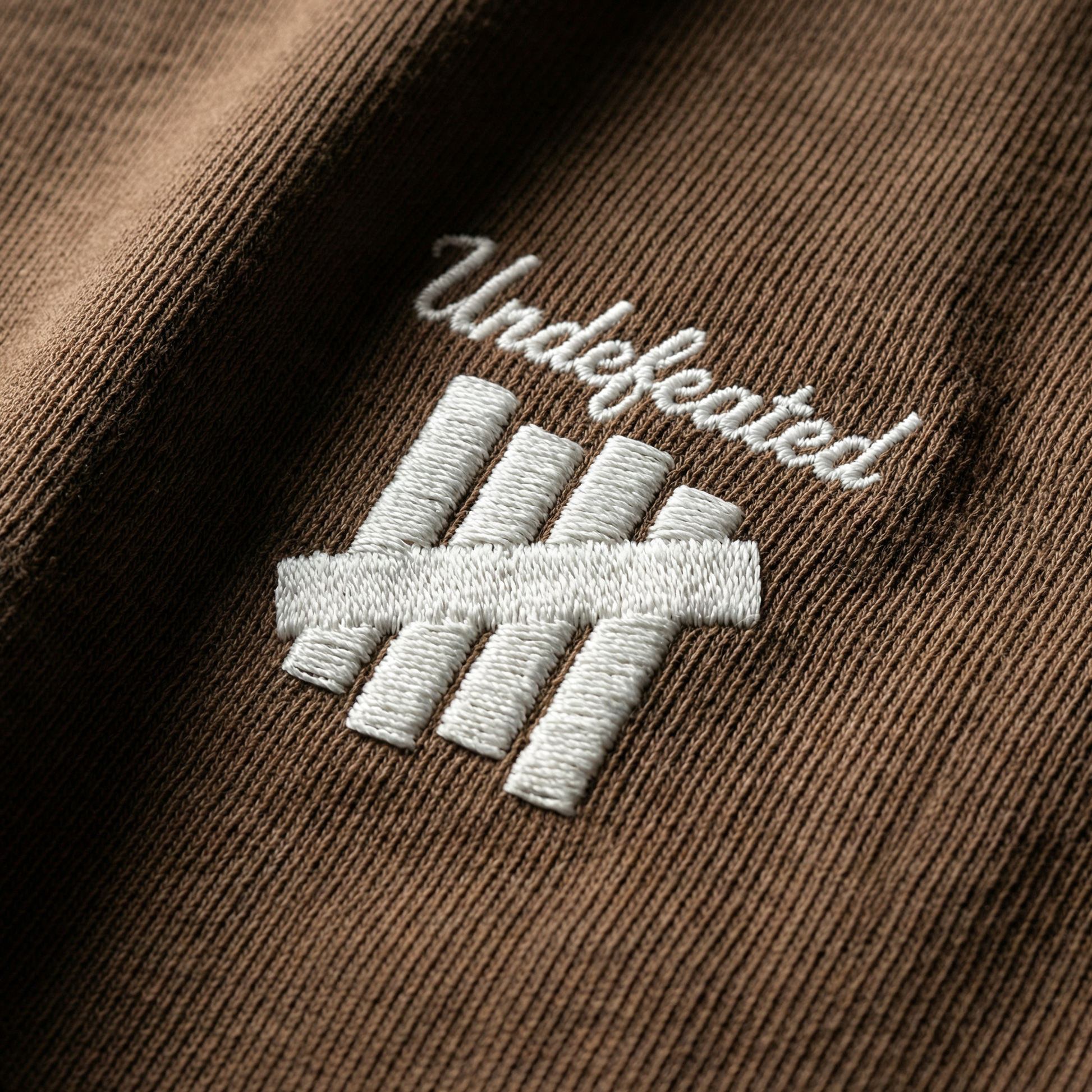 Macro detail of embroidered 4-bar emblem on Brown cotton tee.
