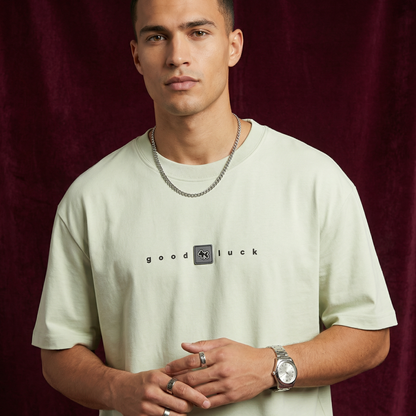 Close-up of WEARON Good Luck Tee in Sage with chest 'good luck' typography, burgundy velvet backdrop.