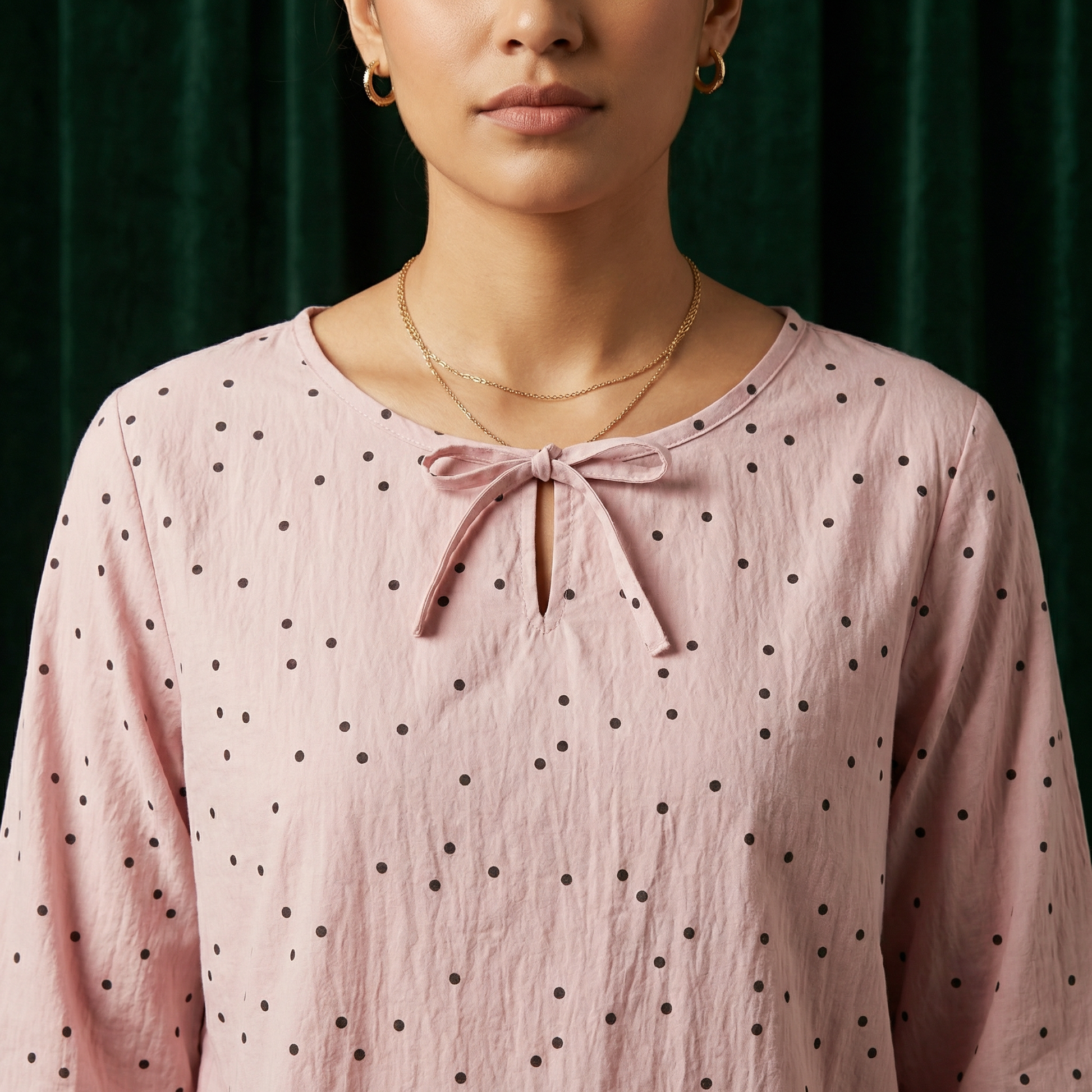 Close-up of WEARON Ribbon-Tie Dot Blouse in Rose with self-tie ribbon at keyhole neckline, deep green velvet backdrop.