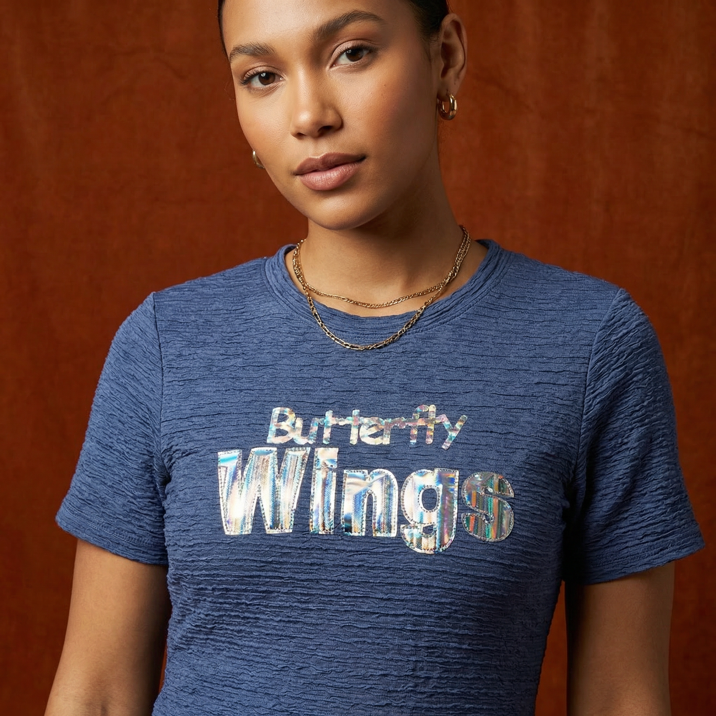 Close-up of WEARON Iridescent Slogan Mini Dress in Navy with iridescent Butterfly Wings graphic, terracotta backdrop.